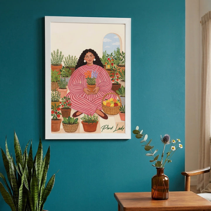Garden Of Her Own Wall Art Frame - framed illustration of a woman in a pink striped outfit surrounded by potted plants, displayed on a teal wall above a wooden table with a vase